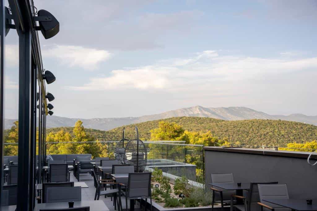 Outdoor terrace dining area with dark furniture overlooking forested hills and mountains under a cloudy sky.