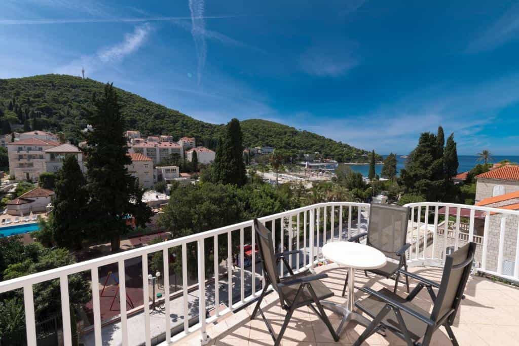 Balcony with table and chairs overlooking coastal town, lush green hills, and blue sea.