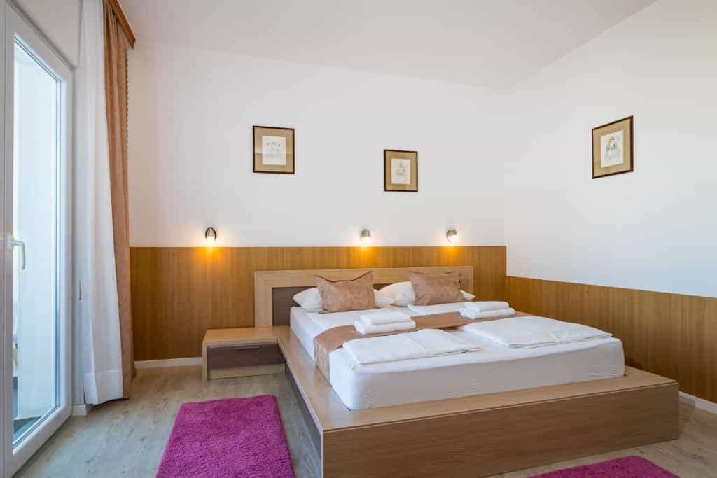 Hotel bedroom with wooden platform bed, white linens, and bright pink rug near balcony door.