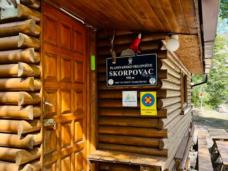Log cabin entrance with "Skorpovac" sign, wooden door, and outdoor seating in a wooded area.