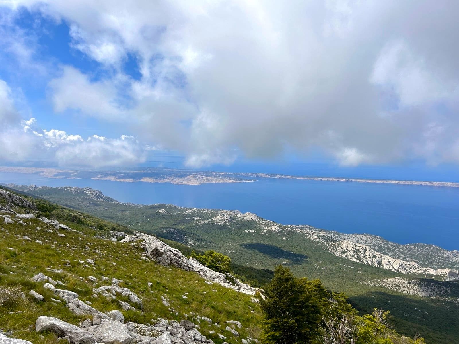 Mountain slope with rocks and green grass overlooking blue sea and distant islands under cloudy sky.