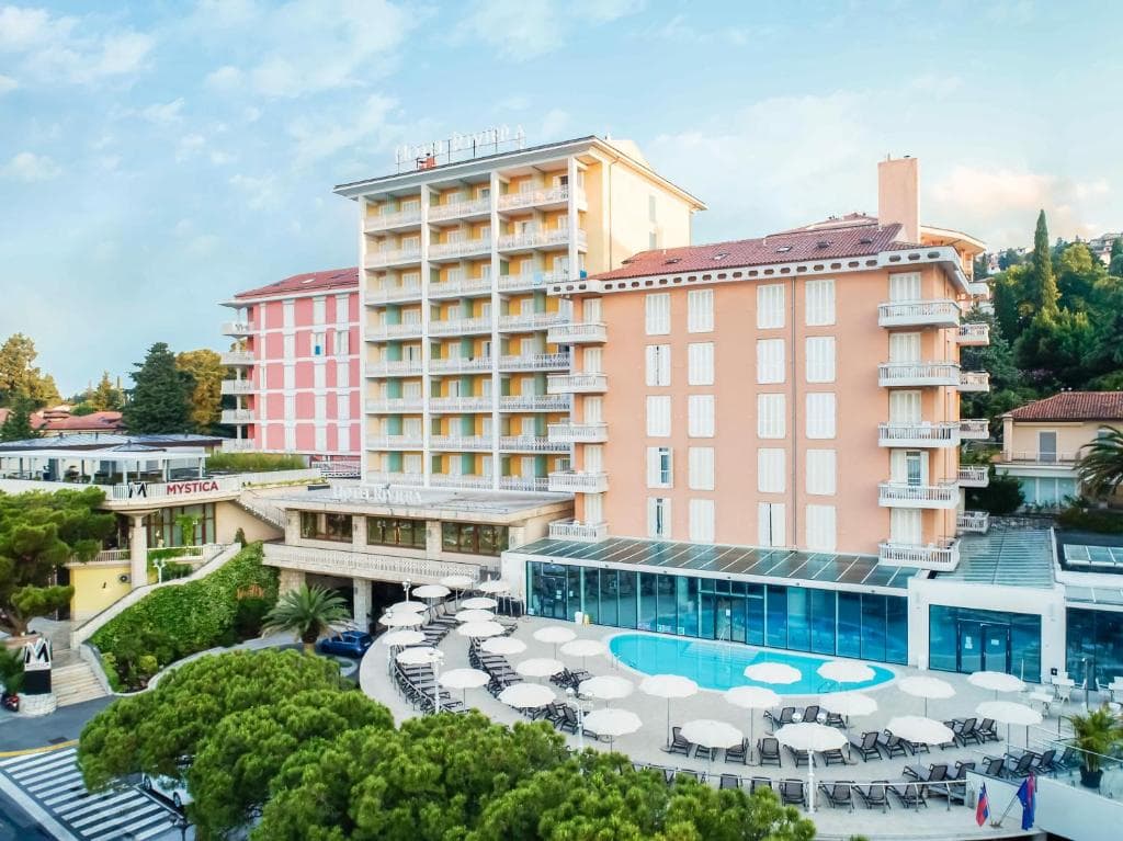 Hotel Riviera building with outdoor pool, lounge chairs, and umbrellas surrounded by greenery.
