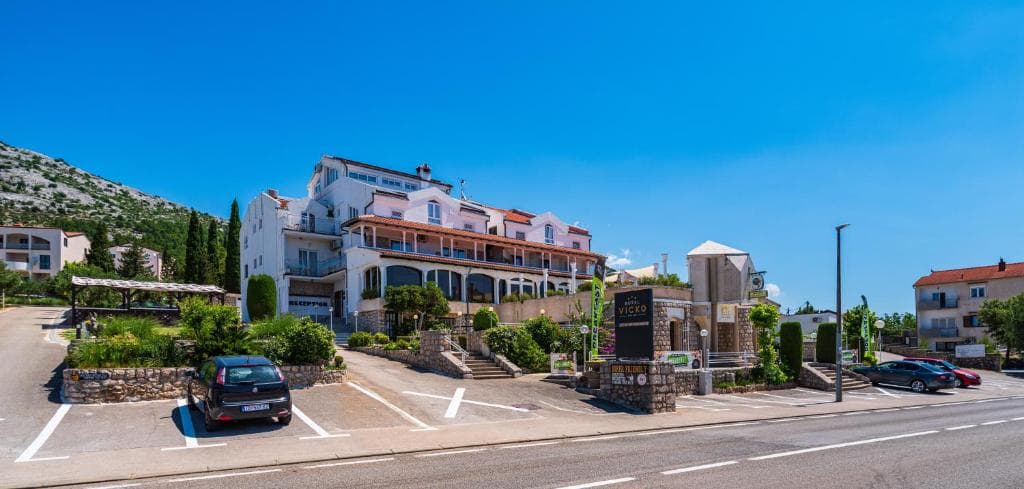 White Hotel Vicko building with parking lot and rocky hillside under clear blue sky.