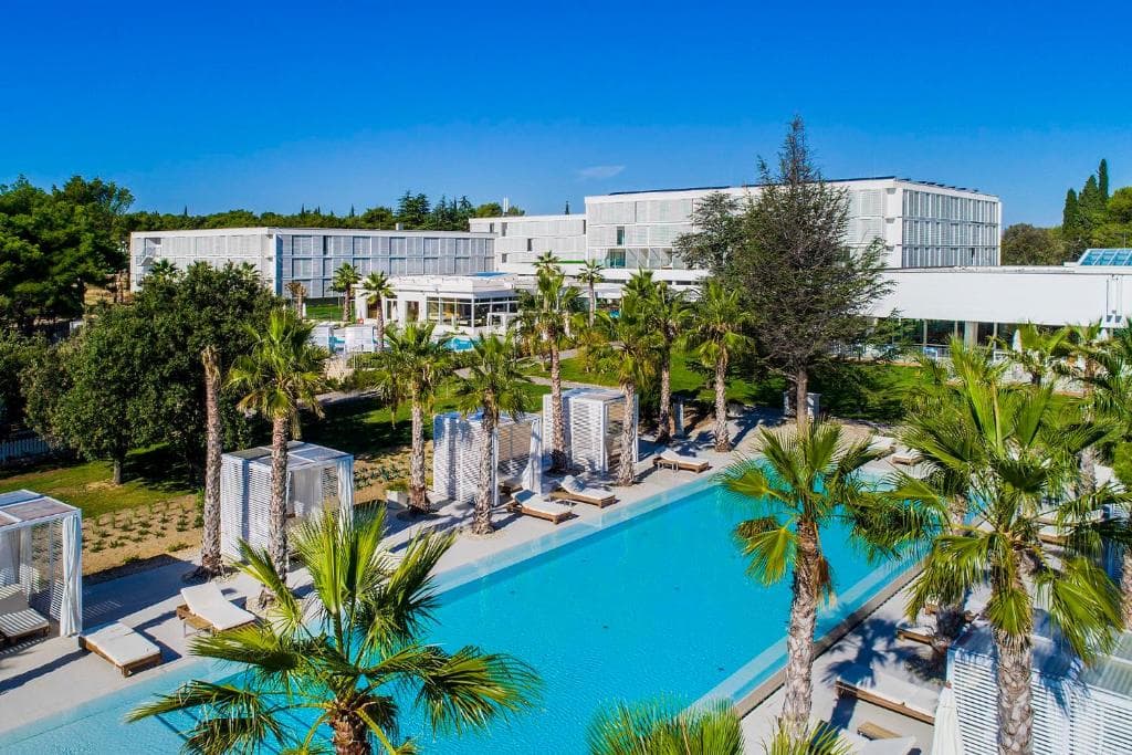 Amadria Park Beach Hotel Jure pool area with palm trees, white cabanas, and modern white hotel buildings.
