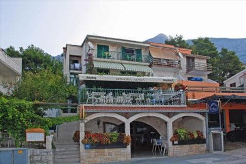 Restaurant Konoba Adria with outdoor seating below mountains, likely in a coastal town.