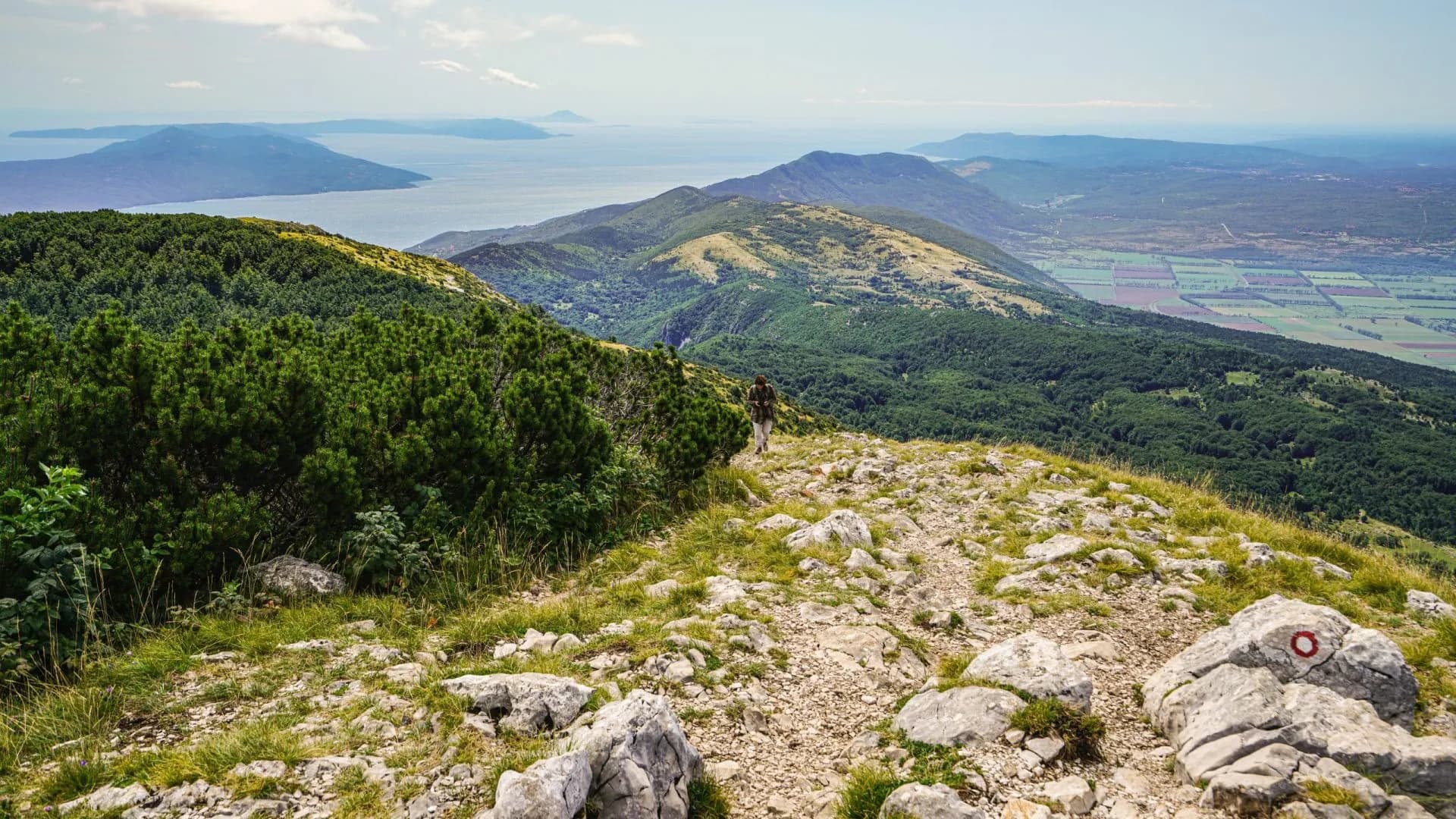 Hiker on rocky mountain trail overlooking forested hills, sea, and distant islands