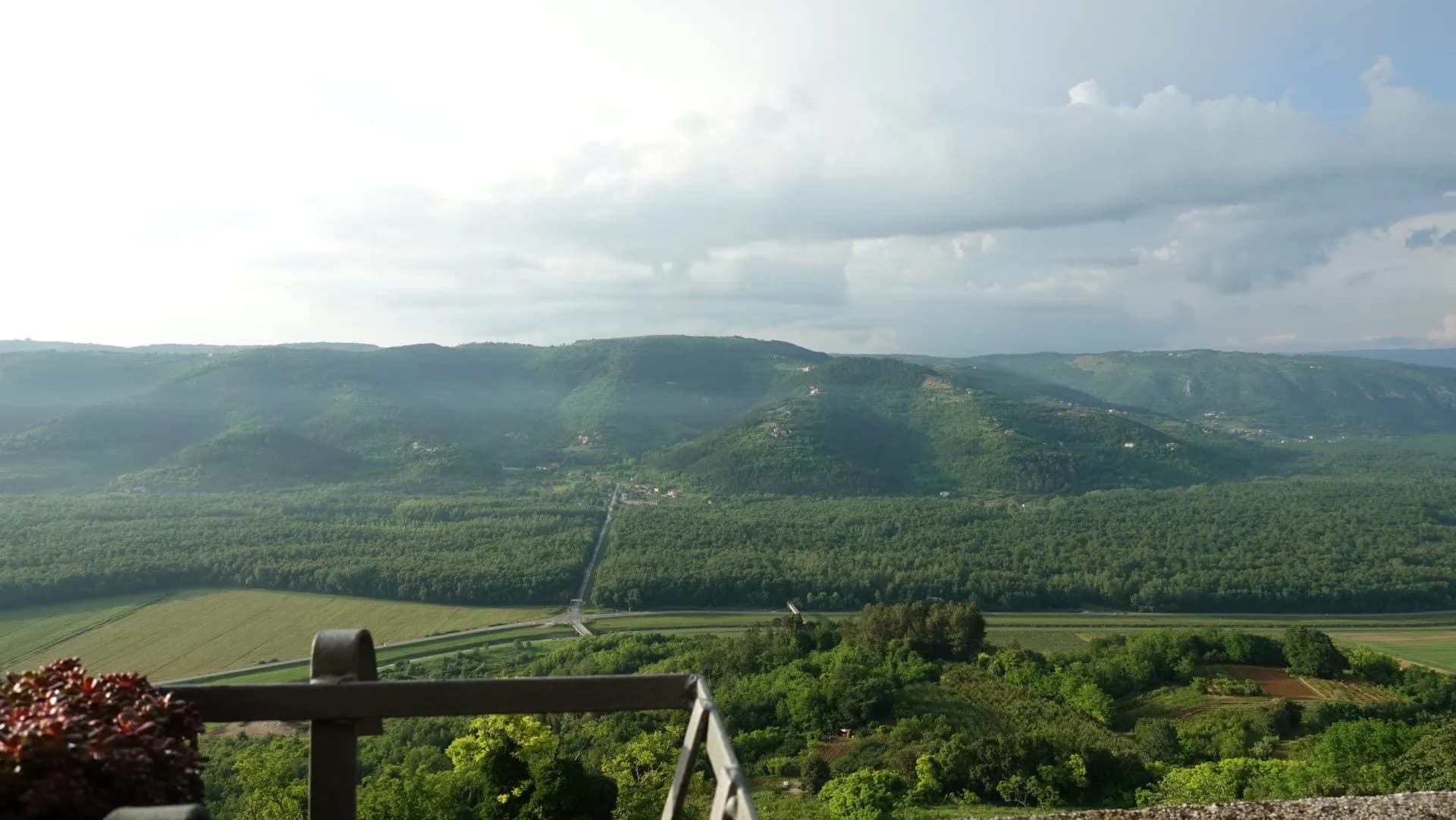 View over green rolling hills, forest, and valley road from a balcony railing.
