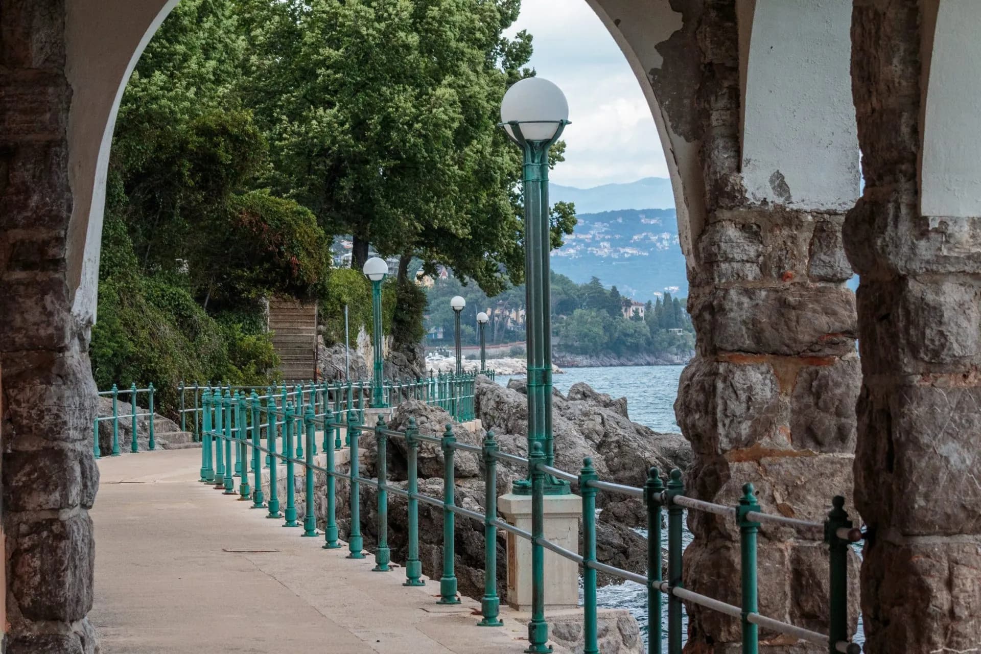 Coastal walkway with stone arches, green railings, and view of sea and distant hillside town.