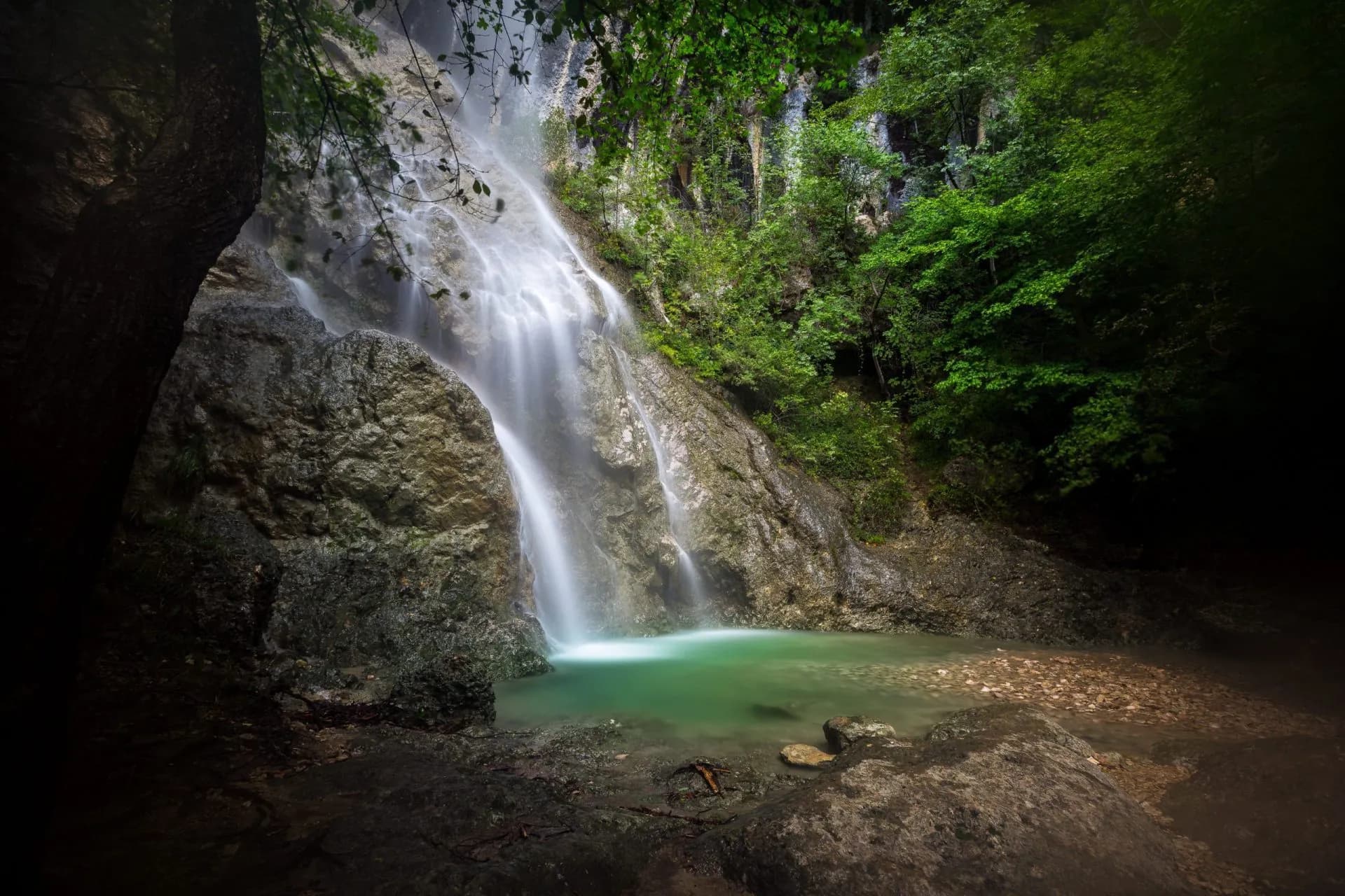 Waterfall cascading over rocks into a green pool surrounded by dense forest foliage.