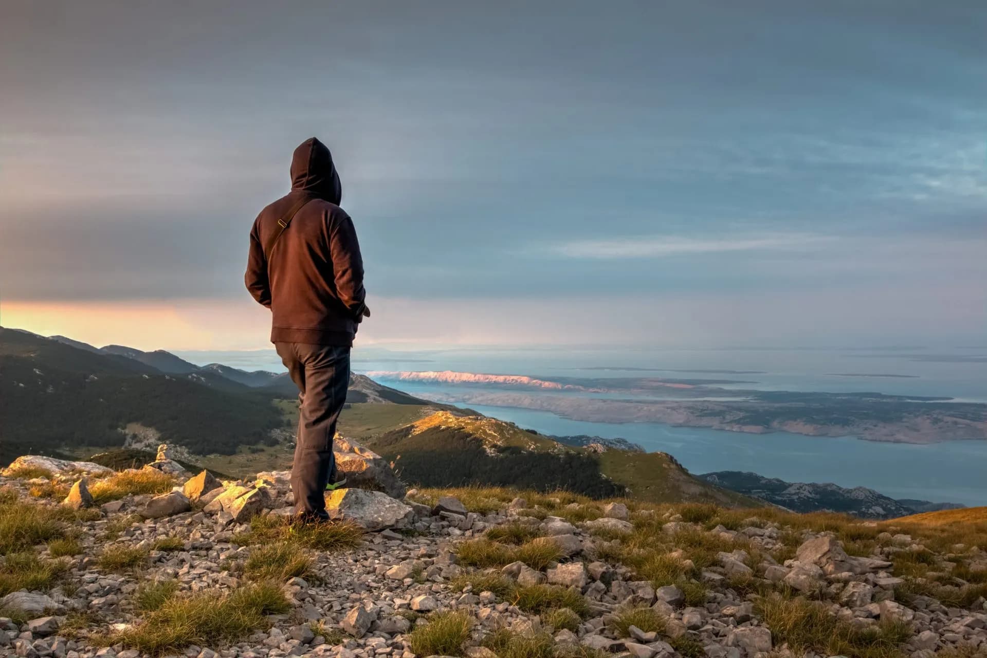 Hiker in hoodie looking over rocky peak at sea and distant islands at sunrise or sunset.