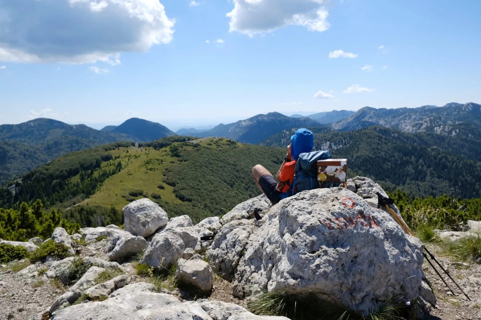Hiker resting with backpack on rocky summit overlooking lush green mountains under blue sky in Croatia.