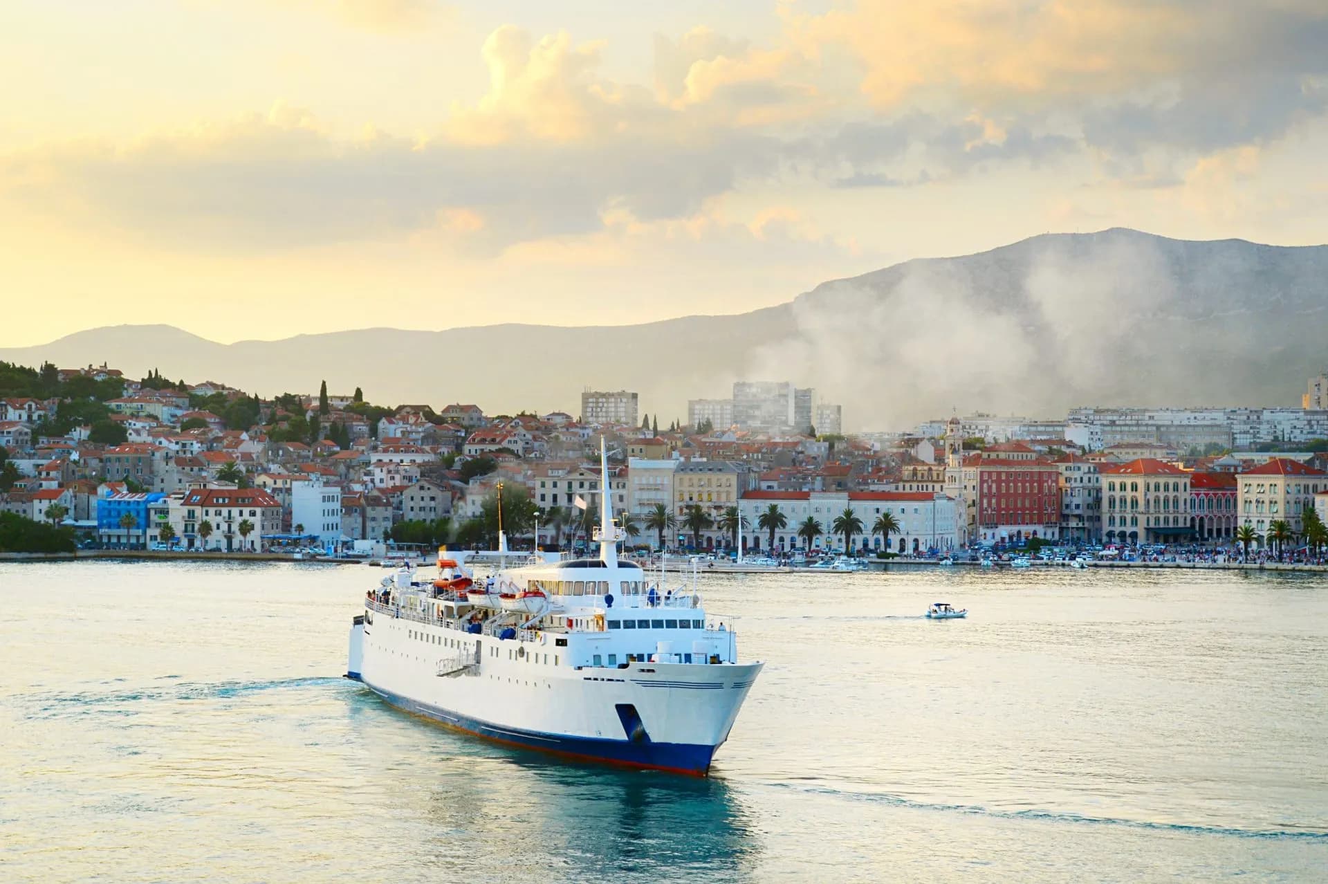Large white ferry boat sailing past a coastal city with red roofs and mountains at sunset.