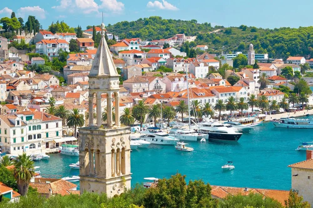 Hvar town harbor with bell tower, white buildings, palm trees, and yachts on turquoise water.