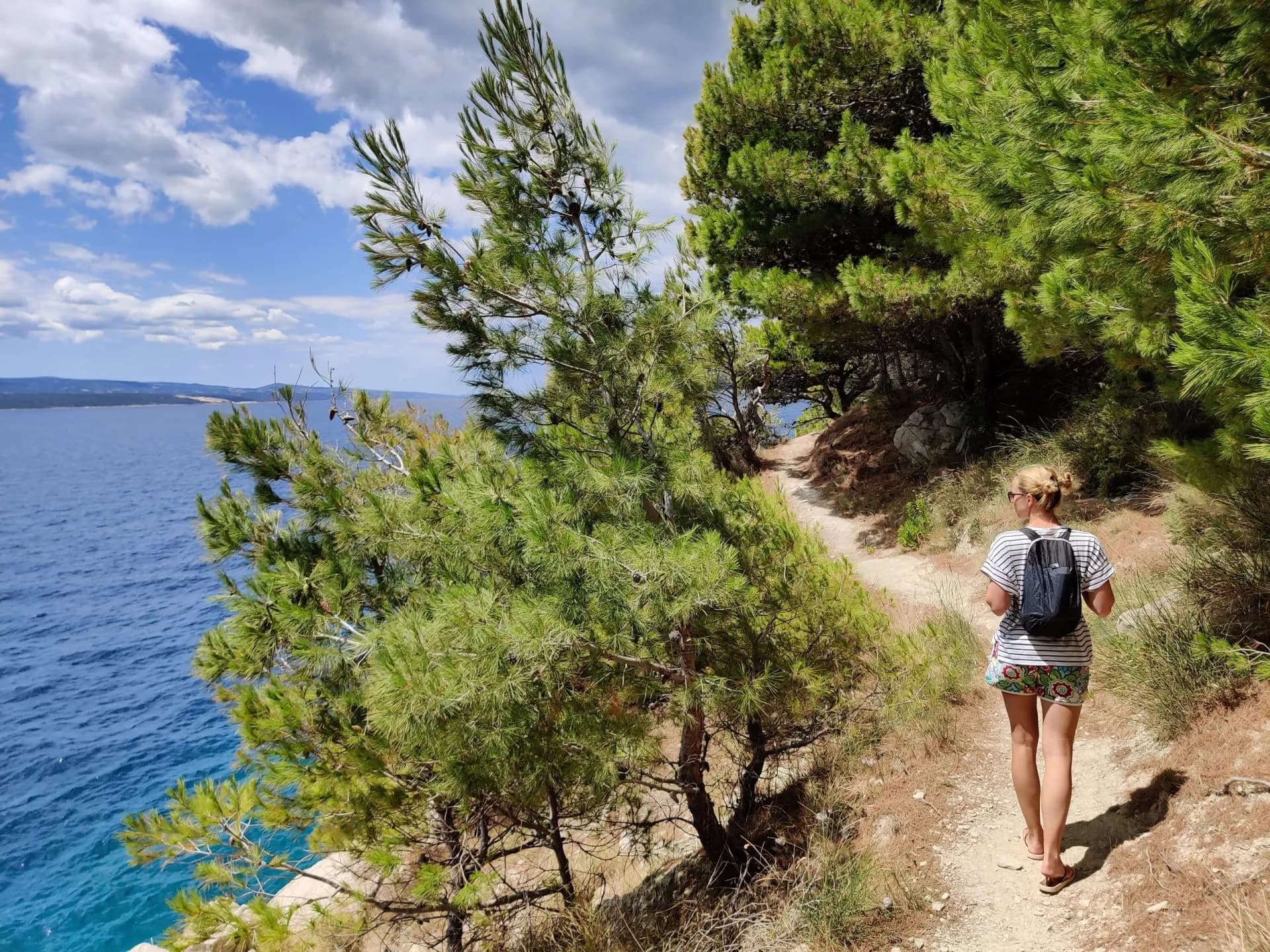 Woman hiking on coastal path overlooking blue sea and distant islands under cloudy sky