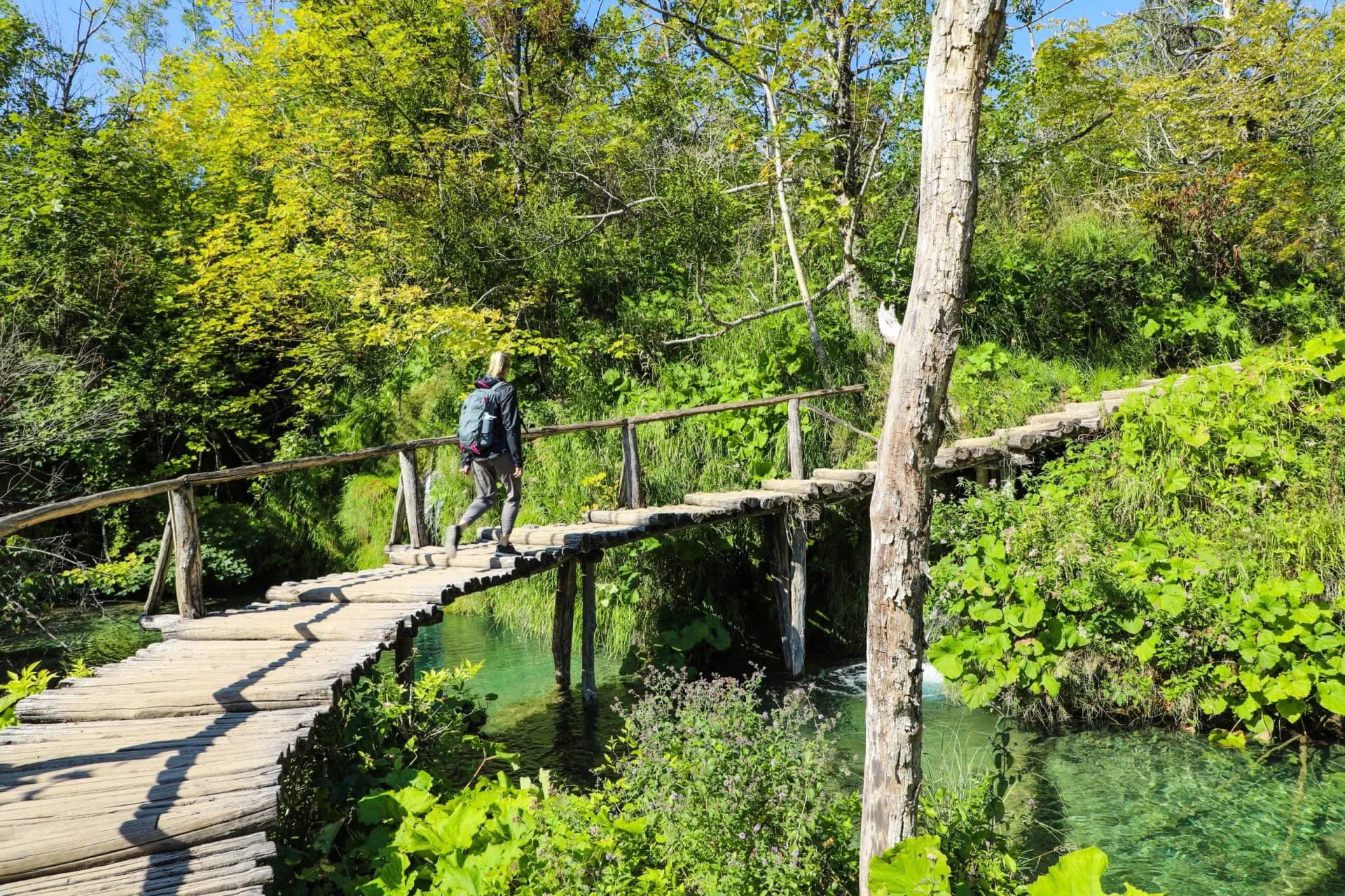 Female hiker with backpack crossing wooden footbridge over turquoise water in Plitvice.