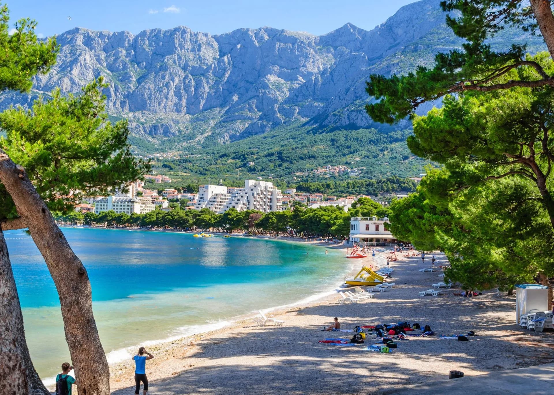 Pebble beach with turquoise water, hotels, and massive mountains on the Makarska coastline.