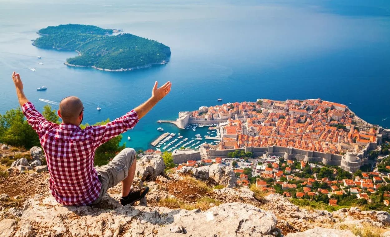 Hiker overlooking walled city of Dubrovnik with orange roofs and Lokrum Island in the Adriatic Sea.