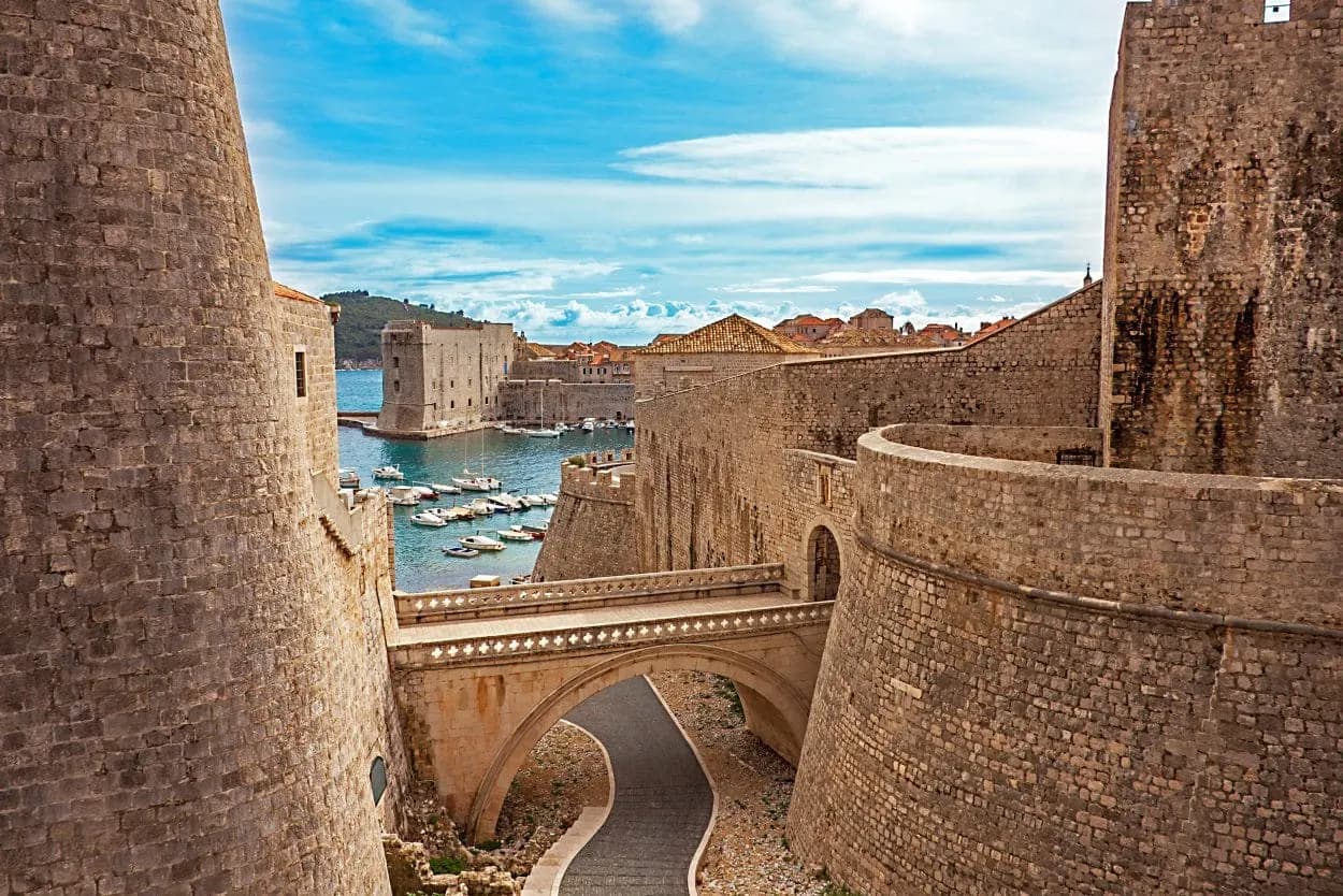 Fort walls of Dubrovnik overlooking harbor with boats and stone bridge under blue sky.