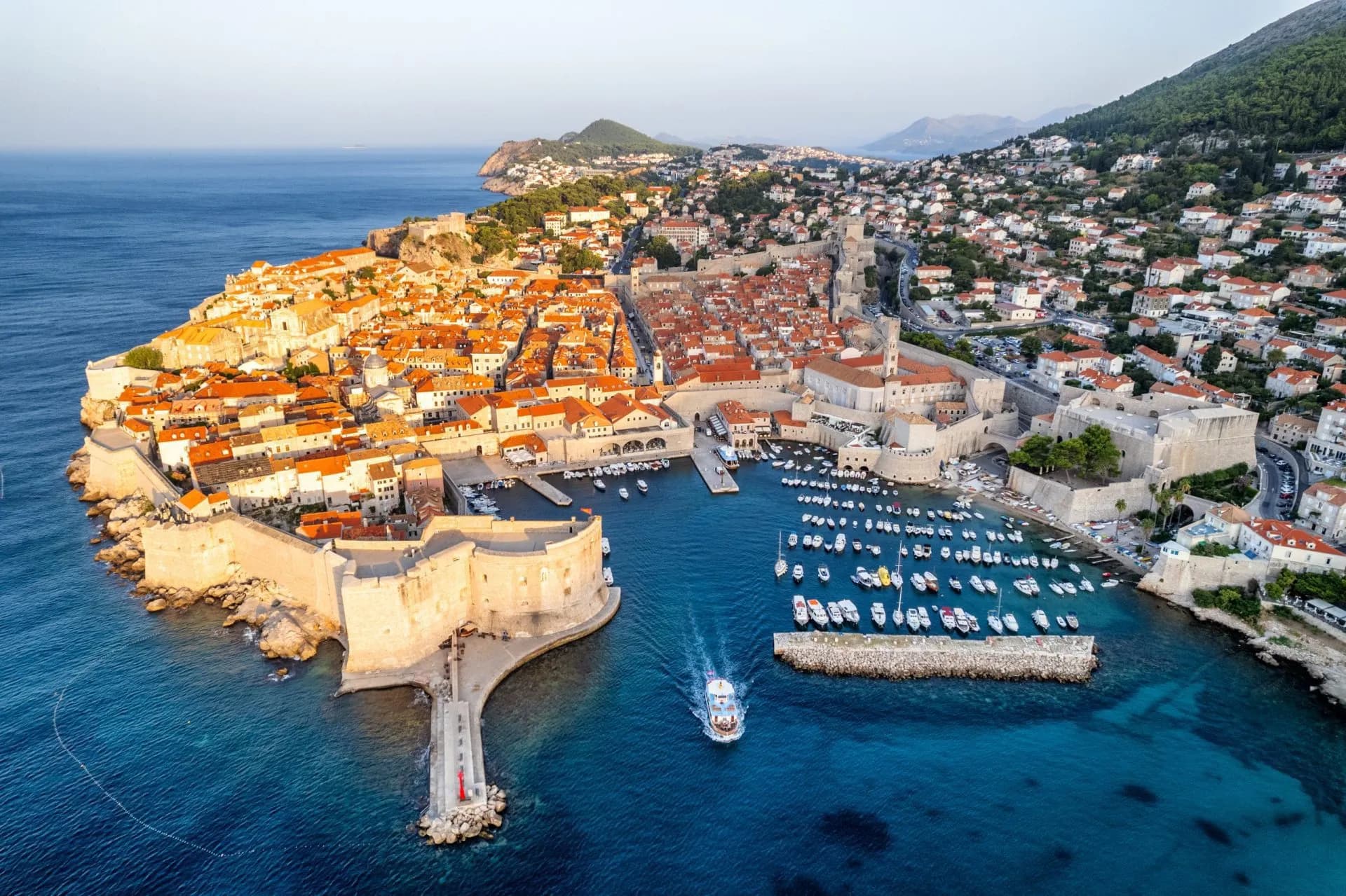 Sunrise over the walled city of Dubrovnik with orange roofs, harbor, and blue Adriatic Sea.