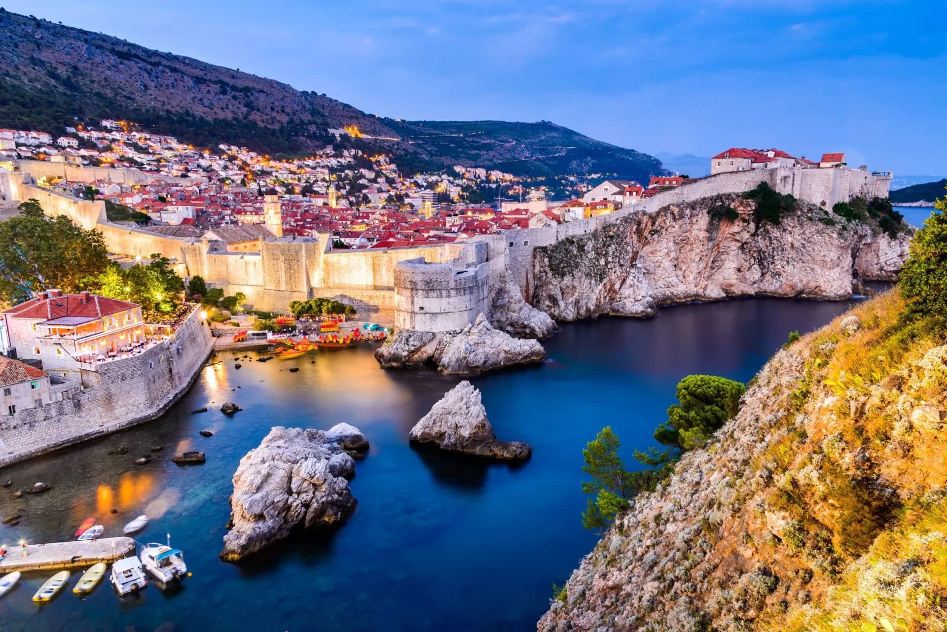 Dubrovnik city walls illuminated at dusk overlooking deep blue cove with small boats and rocky coastline.