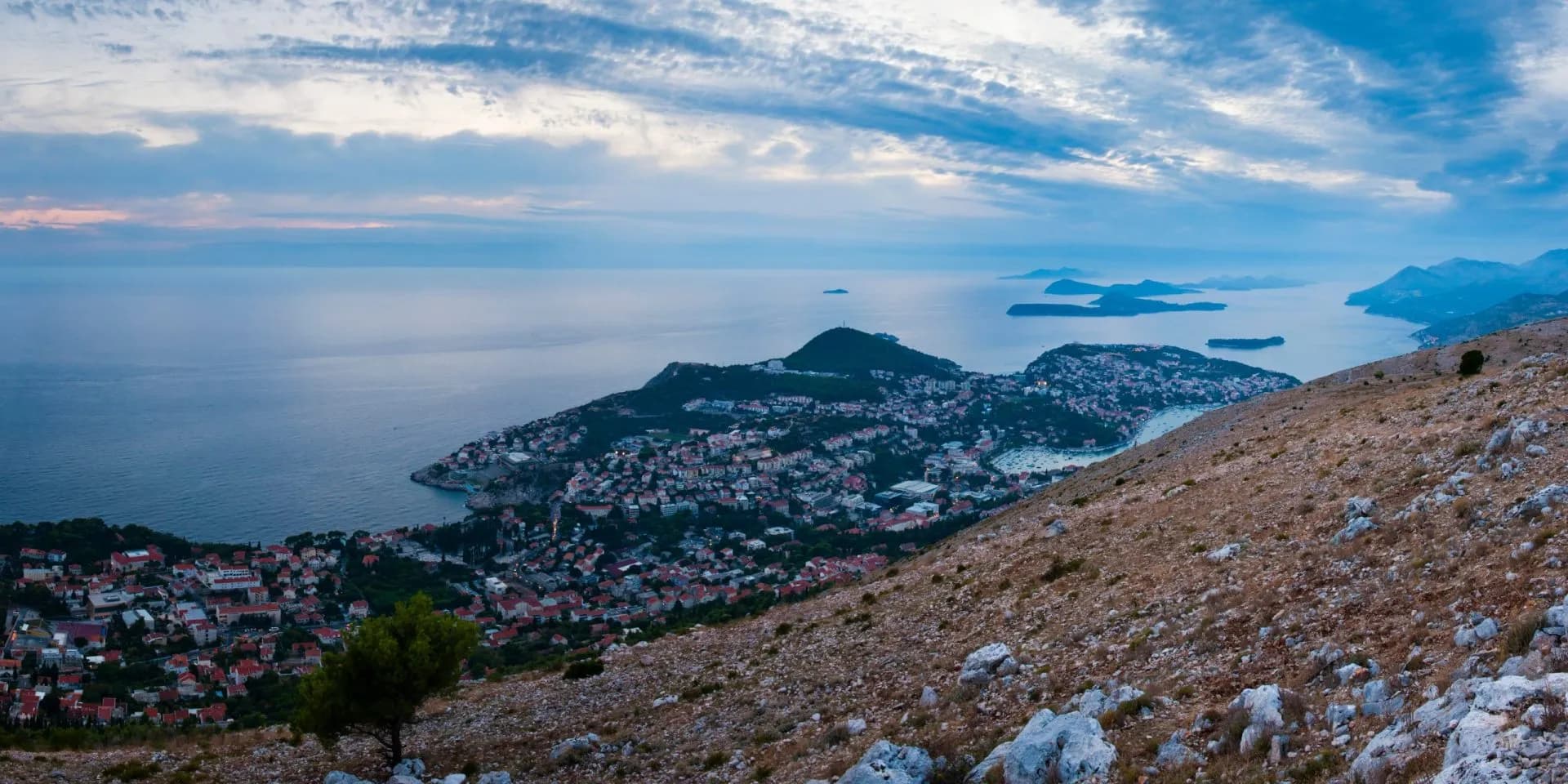 Coastal town nestled by the sea viewed from a rocky, arid hillside under a cloudy sky.