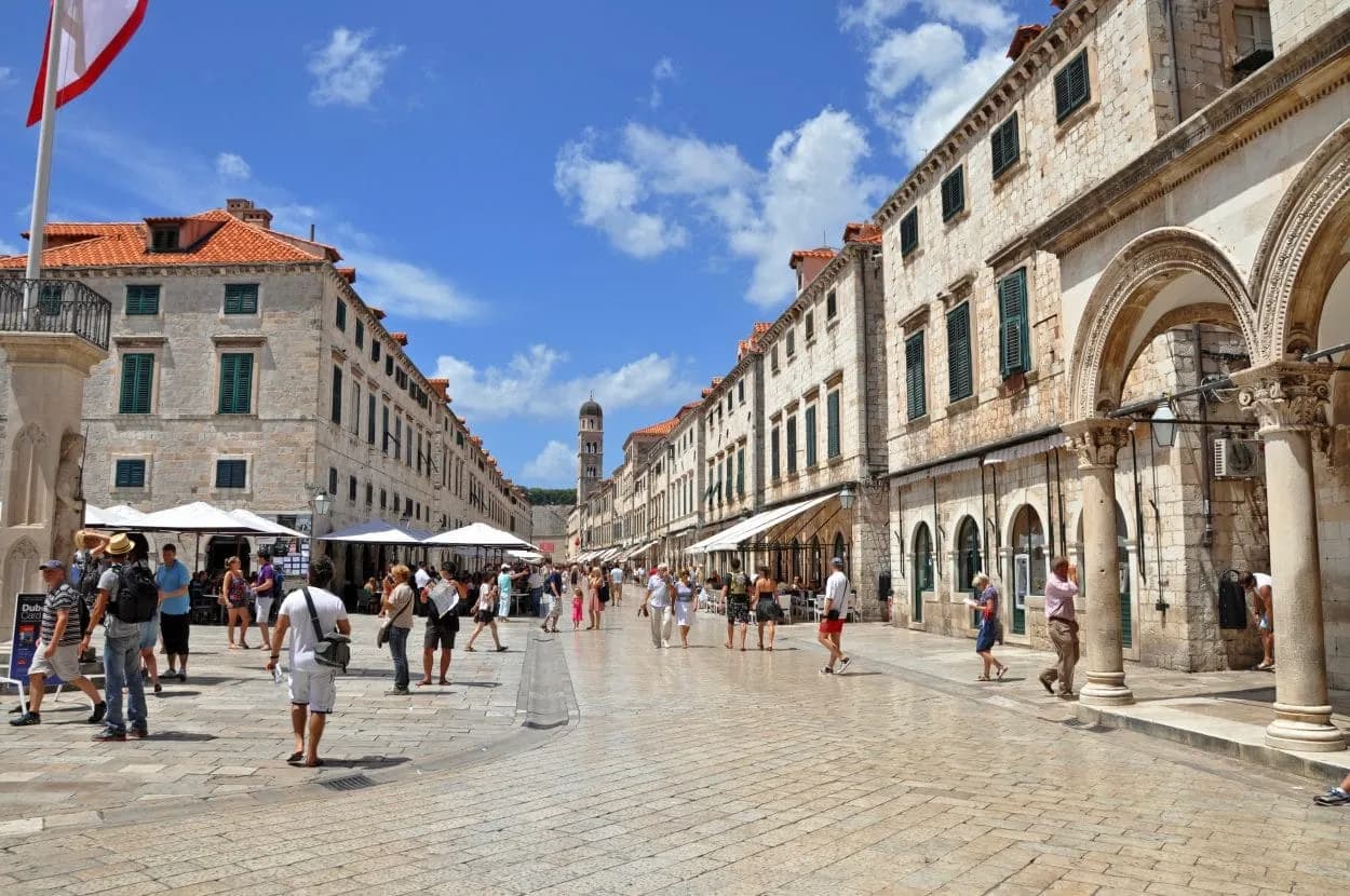 Pedestrians on the main stone street lined with historic buildings in Dubrovnik under a blue sky.