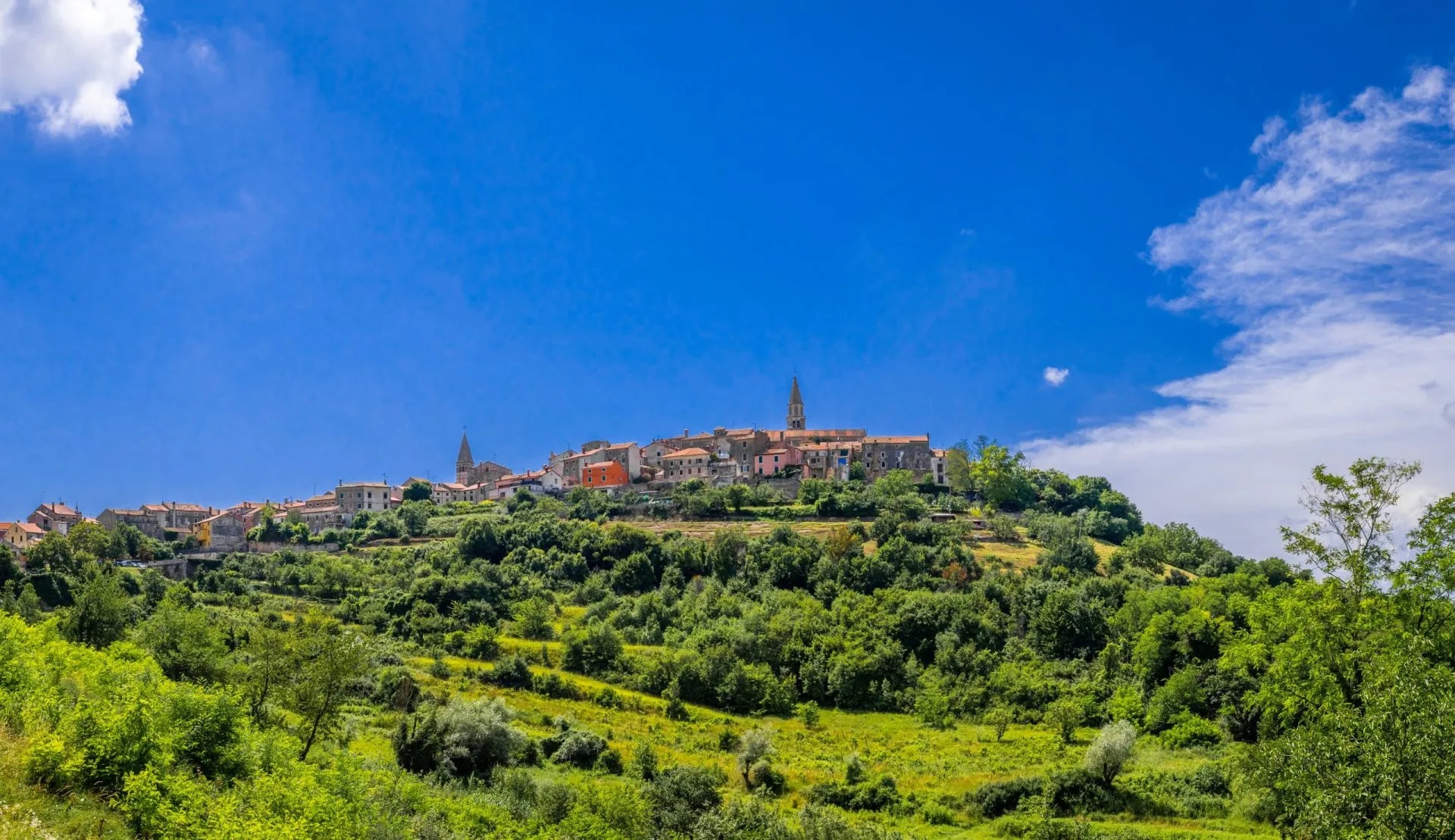 Hilltop village of Buje with terracotta roofs nestled in lush green landscape under a bright blue sky.