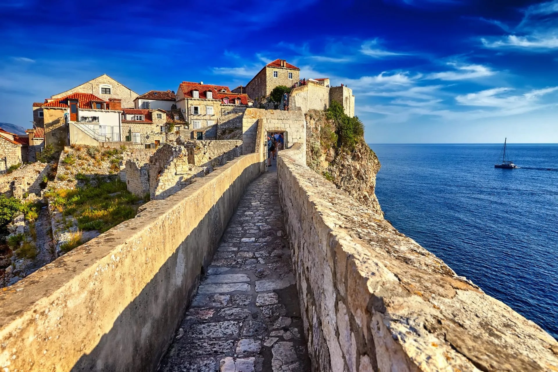 Stone walls of Dubrovnik Old Town overlooking the sea at sunset with a sailboat.
