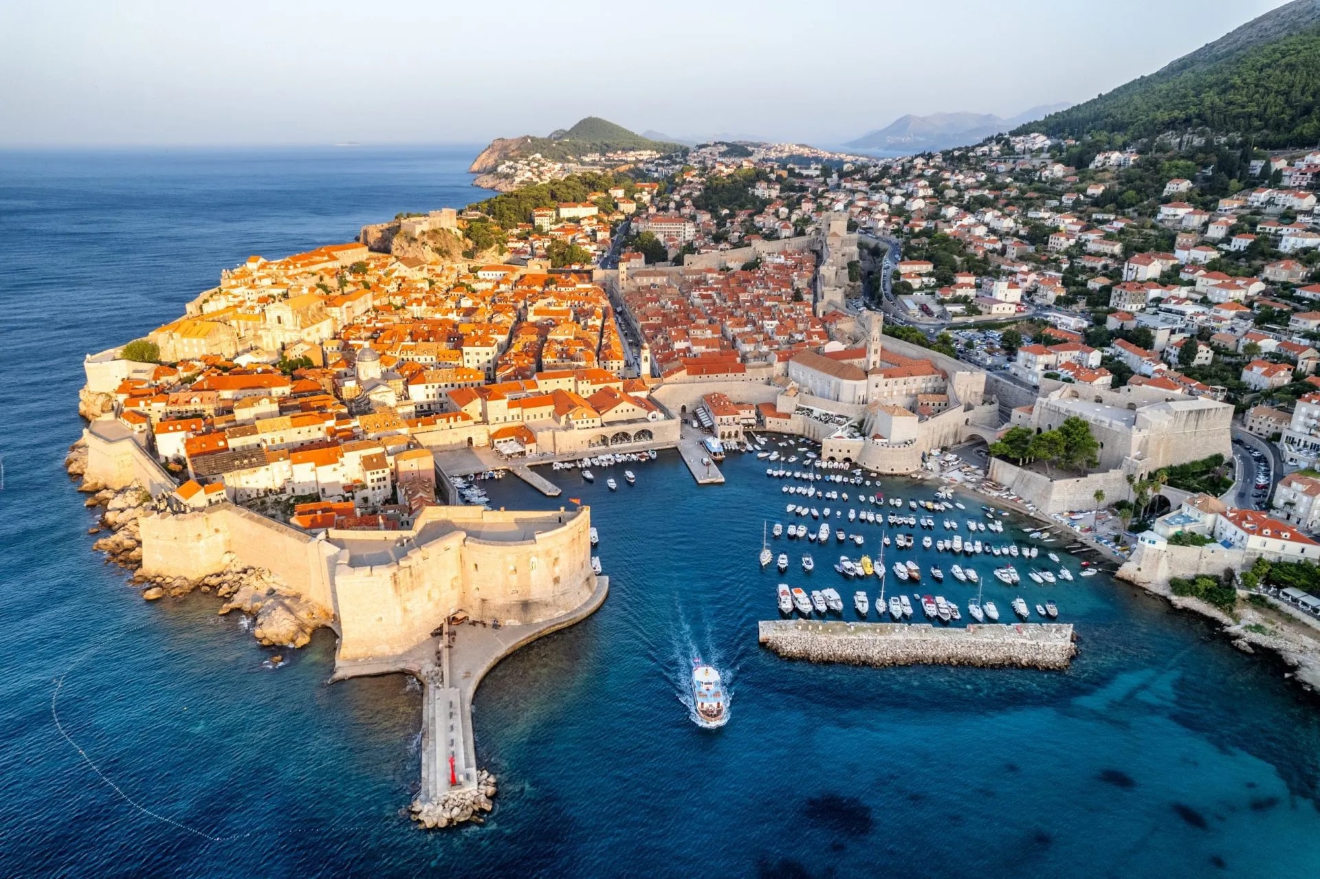 Sunrise over the historic walled city of Dubrovnik with terracotta roofs and a boat leaving the harbor.