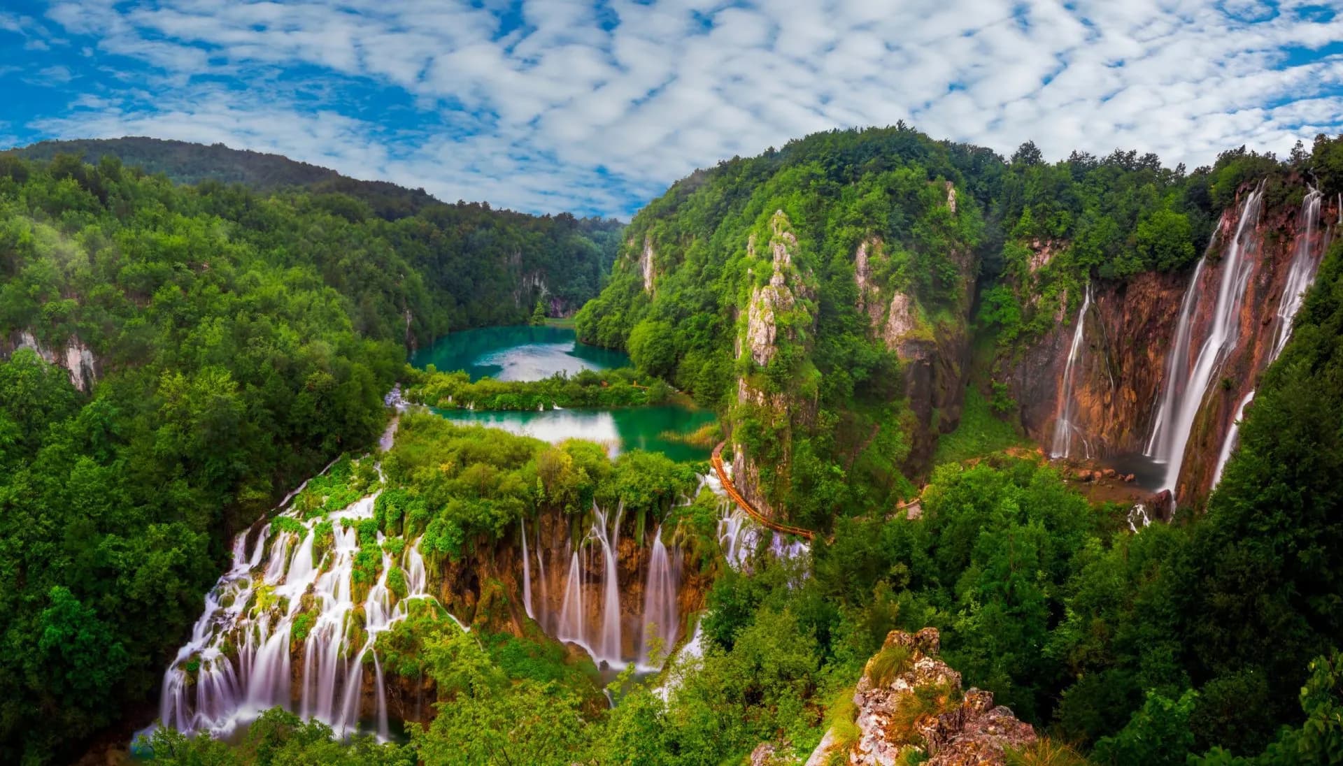 Plitvice Lakes panorama with cascading waterfalls, turquoise pools, and lush green forests under a cloudy blue sky.