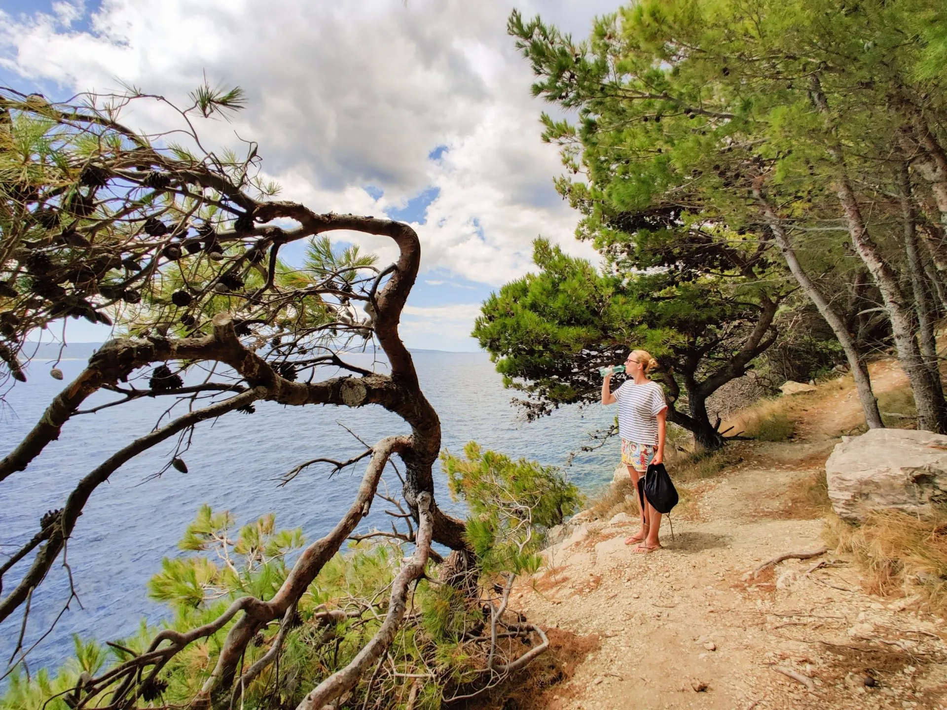 Young active feamle tourist taking a break, drinking water, wearing small backpack while walking on coastal path among pine trees looking for remote cove to swim alone in peace on seaside in Croatia.
